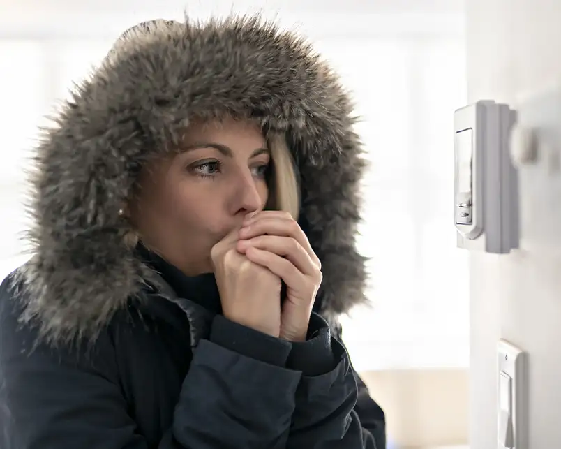 A woman bundled in a winter coat stands near a thermostat inside a cold home, illustrating the need for Precision Climate Control's heating repair services in St. Charles, MO