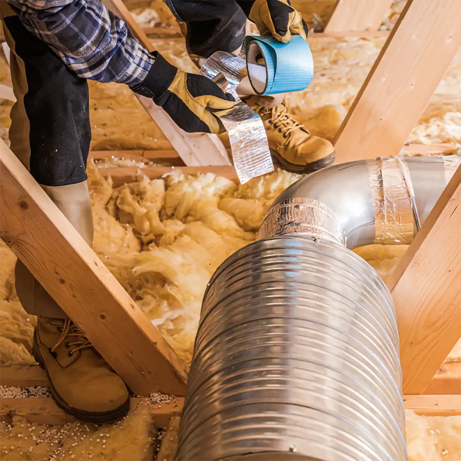 A Precision Climate Control technician in an attic seals flexible silver ductwork with foil tape among fiberglass insulation during a duct installation in St. Charles, MO