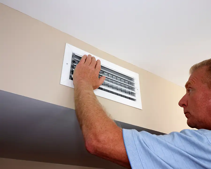 A homeowner reaches up to adjust the louvers on a wall-mounted HVAC supply vent, representing the airflow balancing services offered by Precision Climate Control in St. Charles, MO