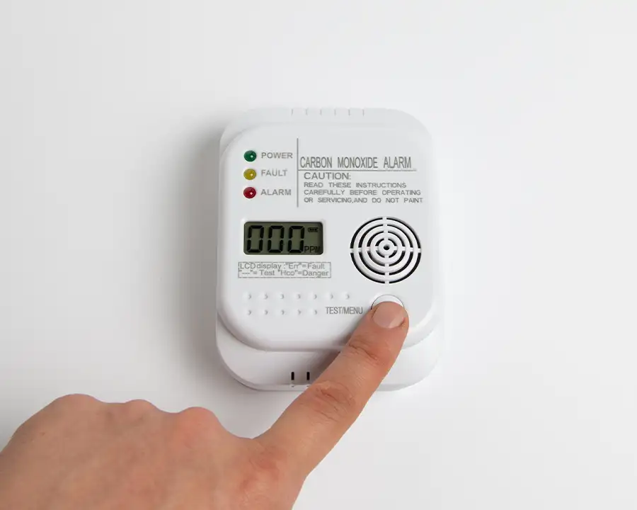 A finger presses the test button on a wall-mounted carbon monoxide alarm with a digital PPM display, highlighting Precision Climate Control's commitment to home safety in St. Charles, MO