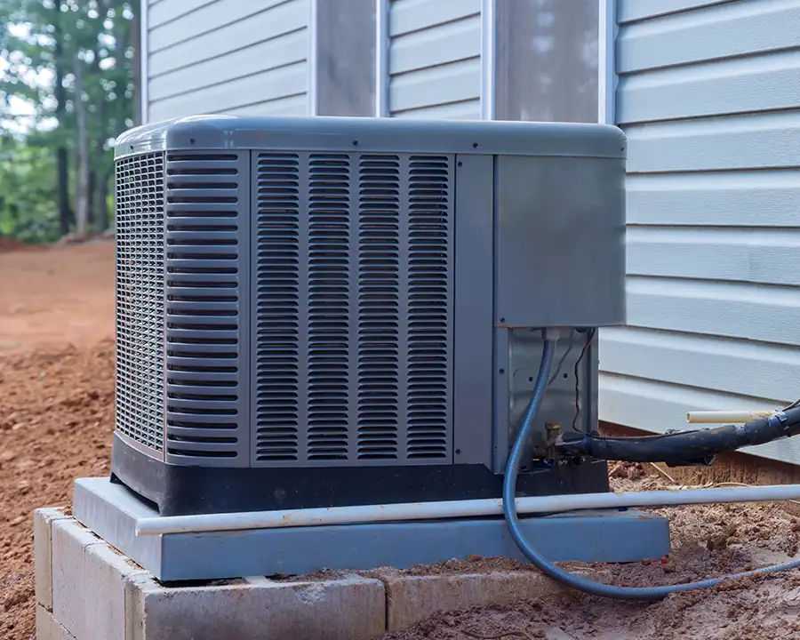 A newly installed gray central air conditioning condenser unit sits on a concrete pad beside a vinyl-sided home surrounded by red clay soil in St. Charles, MO