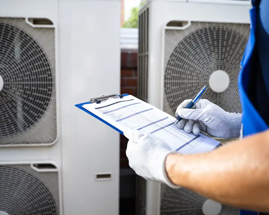 A gloved Precision Climate Control technician records HVAC system data on a blue clipboard while standing in front of multiple outdoor condenser units in St. Charles, MO