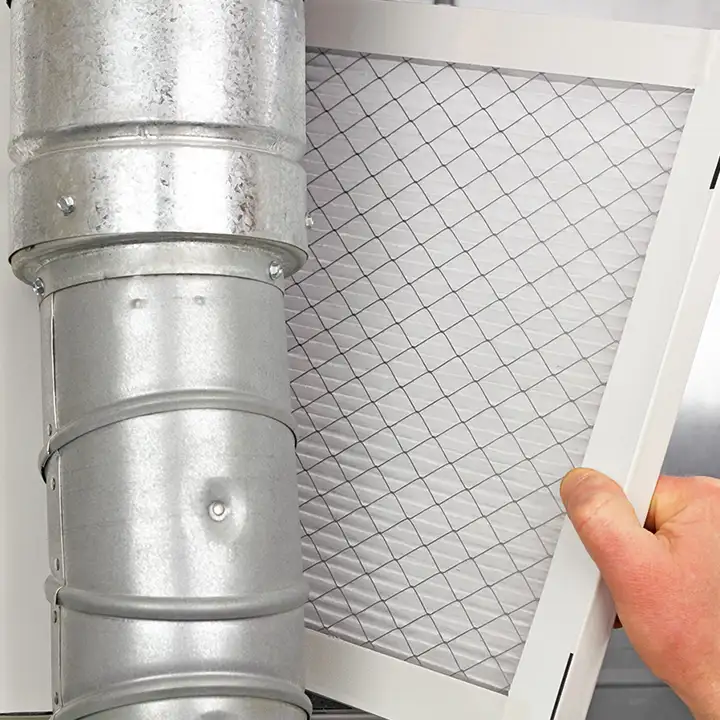 A hand slides a clean white mesh air filter into place beside a round galvanized steel duct connection, representing Precision Climate Control's furnace maintenance and filter replacement services in St. Charles, MO
