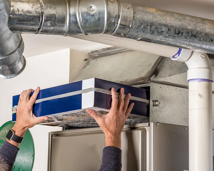 A homeowner slides a large blue high-efficiency air filter into the return air plenum of a furnace system with exposed overhead ductwork, as serviced by Precision Climate Control in St. Charles, MO