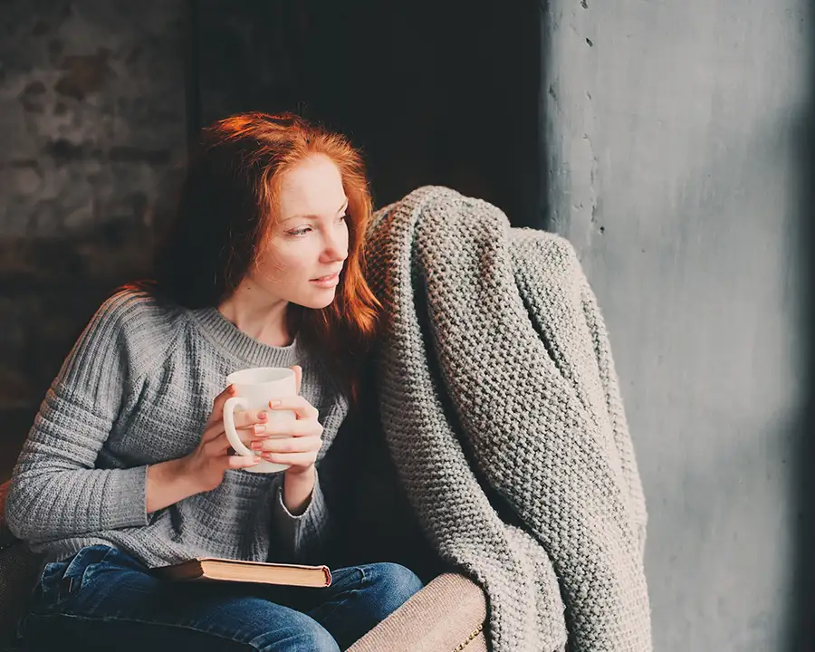 A woman with red hair sits contentedly in a cozy chair with a warm mug and a book, enjoying the comfortable indoor temperature maintained by Precision Climate Control in St. Charles, MO
