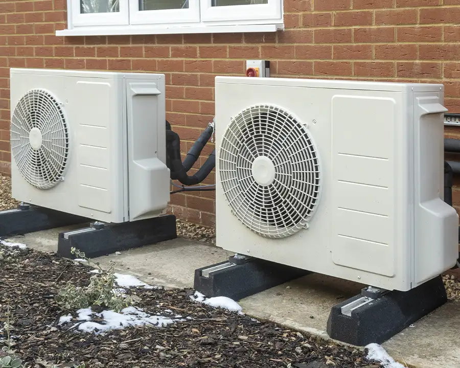 Two white outdoor heat pumps installed side by side on rubber pads against a brick wall with light snow on the ground, representing Precision Climate Control's heating solutions in St. Charles, MO