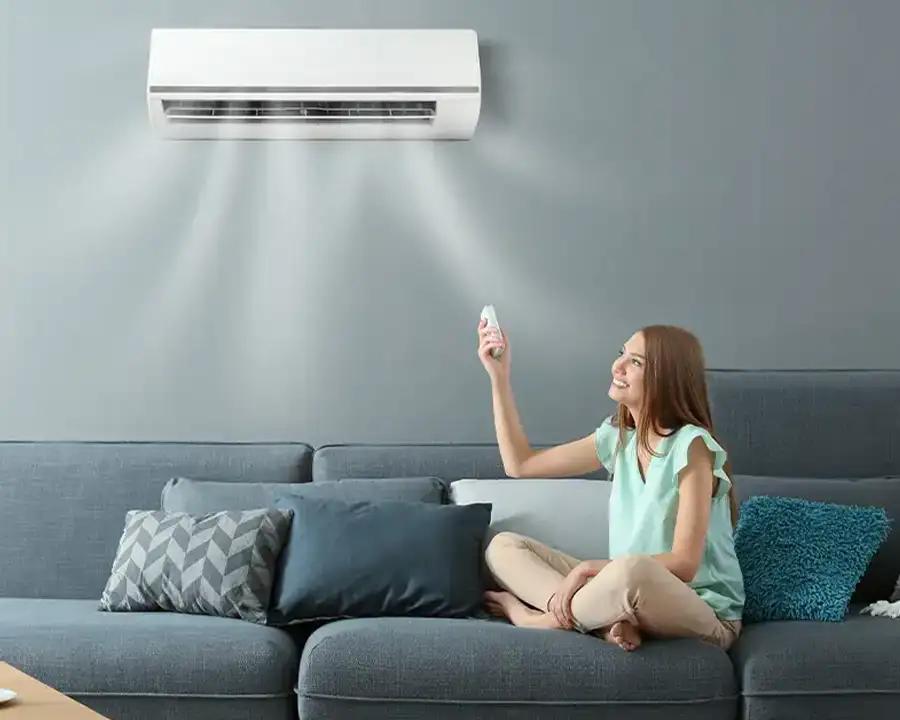 A woman seated cross-legged on a gray sofa points a remote at a wall-mounted ductless mini-split air handler circulating cool air in a modern living room, as installed by Precision Climate Control in St. Charles, MO