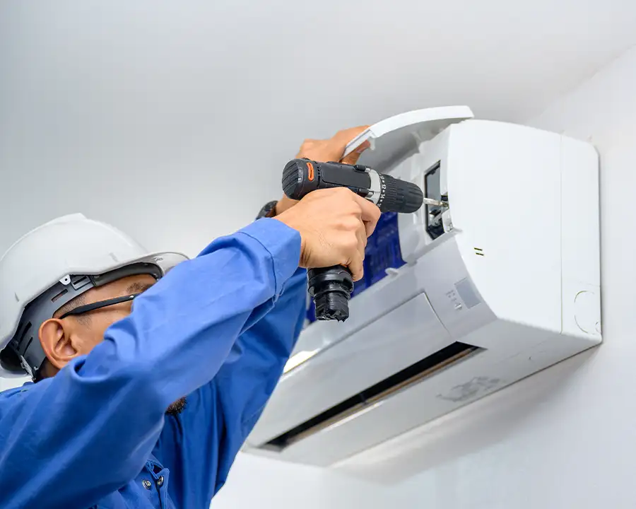 A Precision Climate Control technician in a hard hat and blue jacket uses a power drill to mount the indoor air handler of a ductless mini-split system to a wall in St. Charles, MO