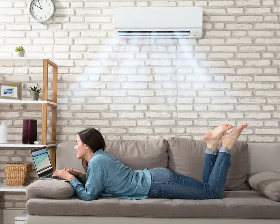 A woman relaxes on a beige couch using her laptop beneath a wall-mounted ductless mini-split blowing cool air, illustrating the home comfort solutions provided by Precision Climate Control in St. Charles, MO