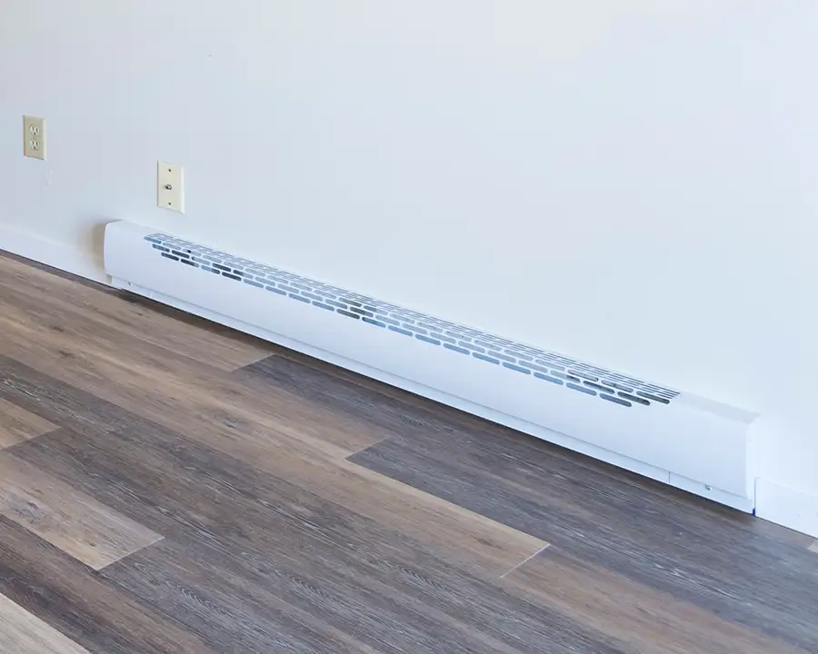 A white electric baseboard heater runs along the base of a white wall above wood-look vinyl flooring, representing supplemental heating options serviced by Precision Climate Control in St. Charles, MO