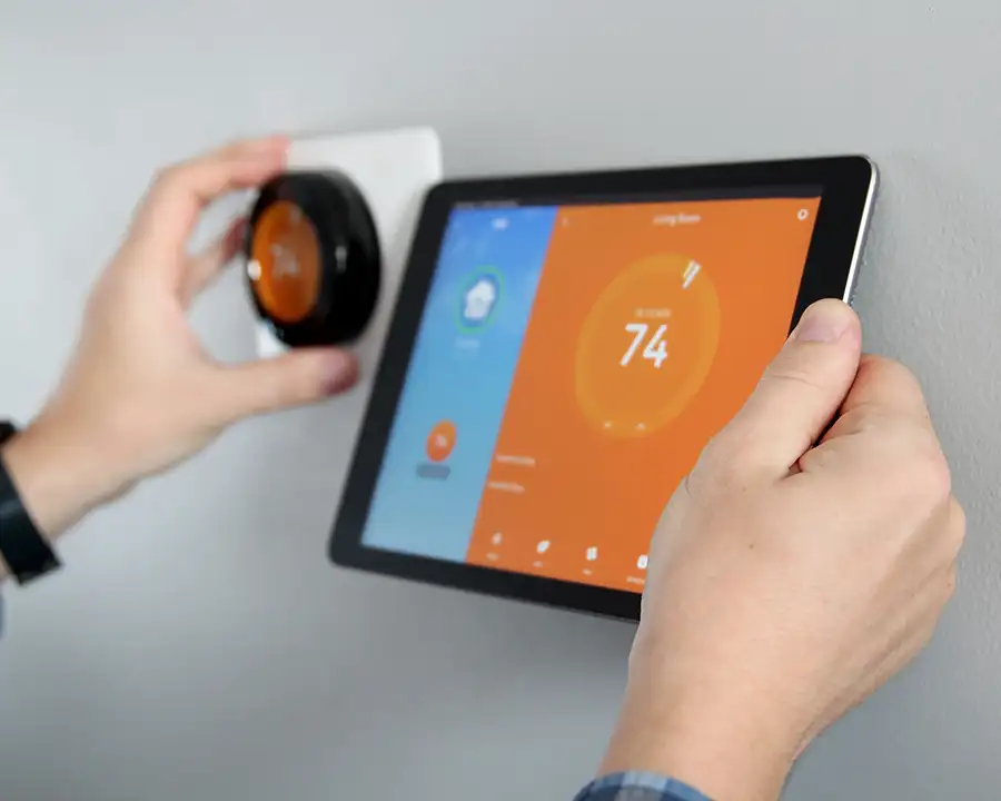 A homeowner adjusts a round smart thermostat on the wall while viewing the corresponding 74-degree temperature reading on a tablet app, representing Precision Climate Control's WiFi thermostat services in St. Charles, MO