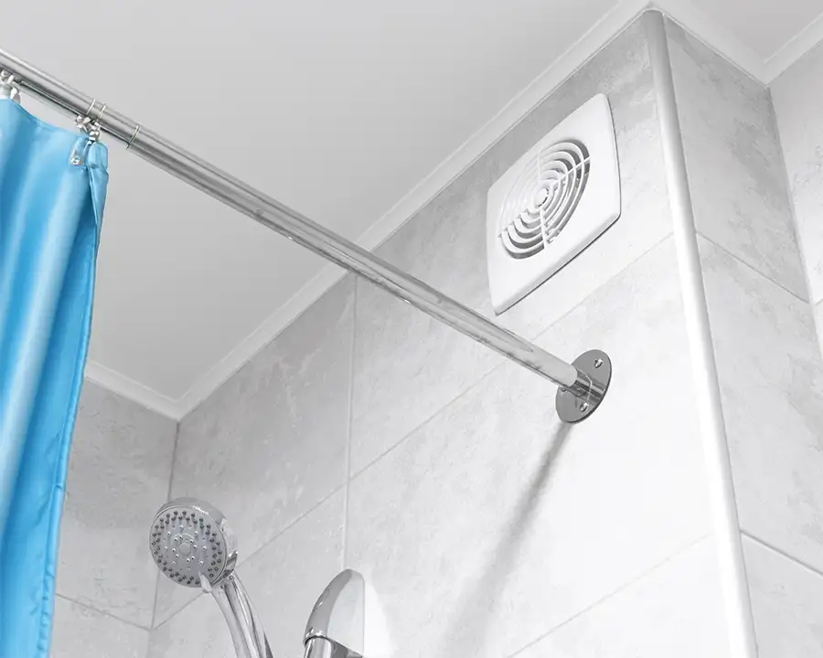 A white bathroom exhaust fan is mounted high on a tiled shower wall beside a blue curtain and chrome showerhead, representing Precision Climate Control's ventilation fan installation services in St. Charles, MO