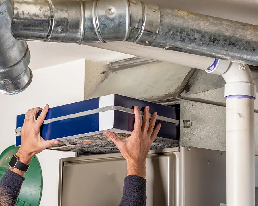 A homeowner pulls a large blue high-efficiency air filter from a furnace return air plenum beneath exposed galvanized ductwork, as serviced by Precision Climate Control in St. Charles, MO