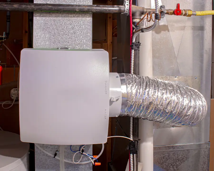 A whole-house humidifier unit with a flexible insulated duct connection is mounted to an HVAC system in a mechanical room, representing Precision Climate Control's indoor humidity control services in St. Charles, MO